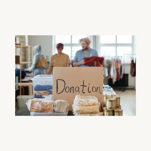 Donation written on a brown cardboard surrounded by food items and three people blurred in the background.
