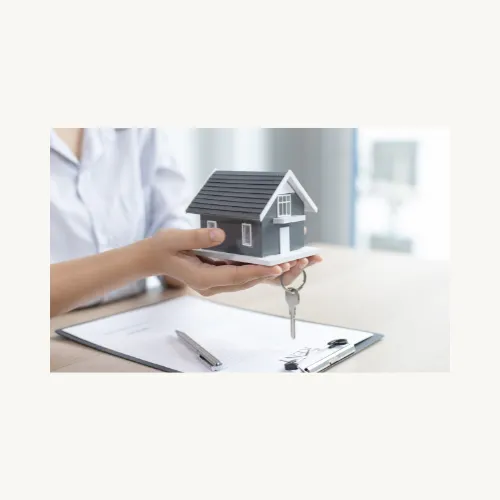 Hands holding a house model with a key between the fingers and a clipboard with a contract laying on a table.